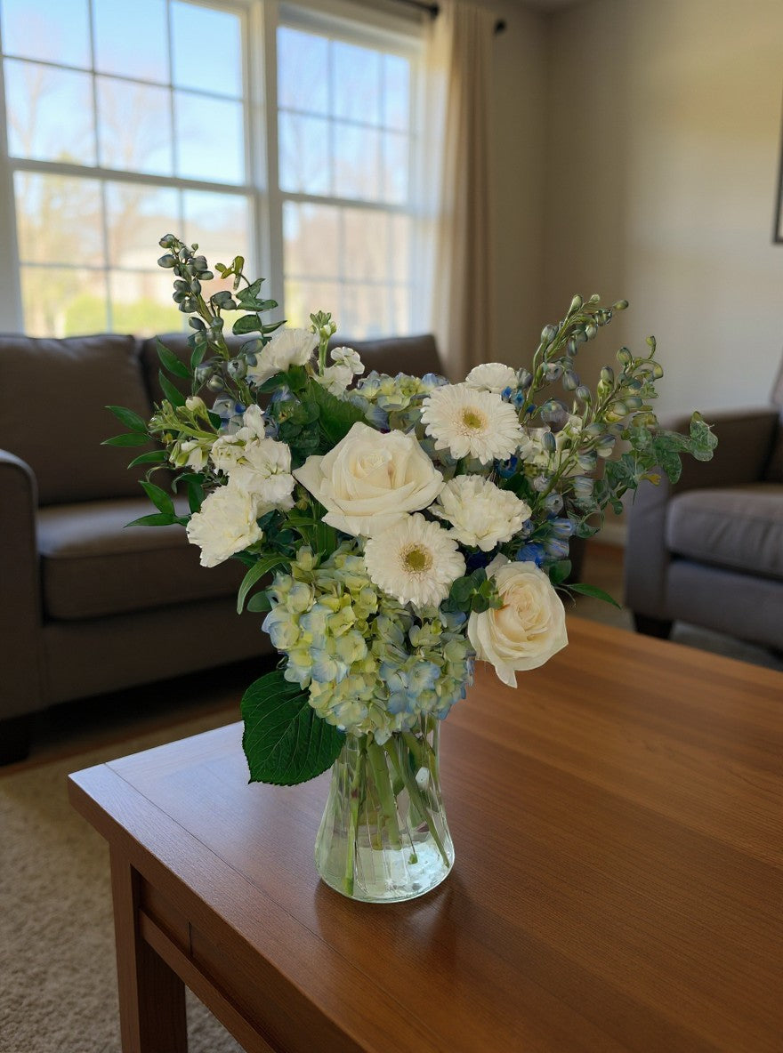 Lakewood Florist Crisp Sky arrangement: white roses, delphiniums, hydrangeas, and greenery in a clear vase.