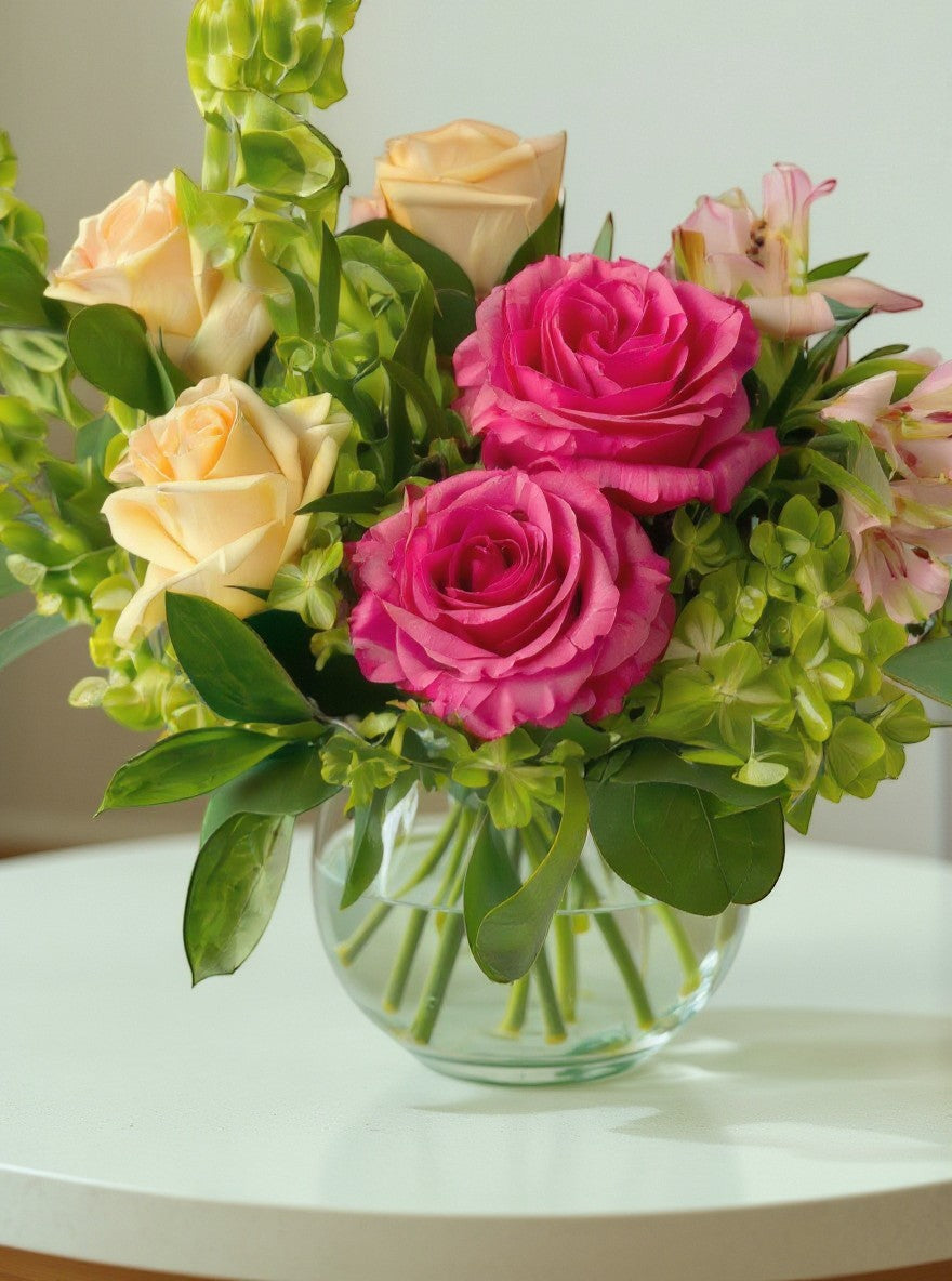 Lakewood Florist bright pink and peach roses with green foliage in a clear glass vase.